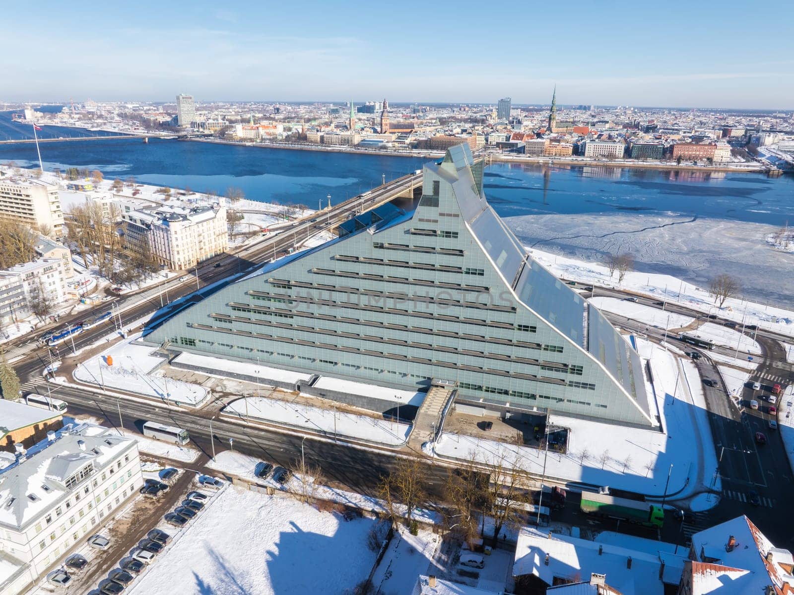 Snow covered National Library of Latvia by the Daugava River with Riga's historic old town spires in the background, showcasing a winter landscape.