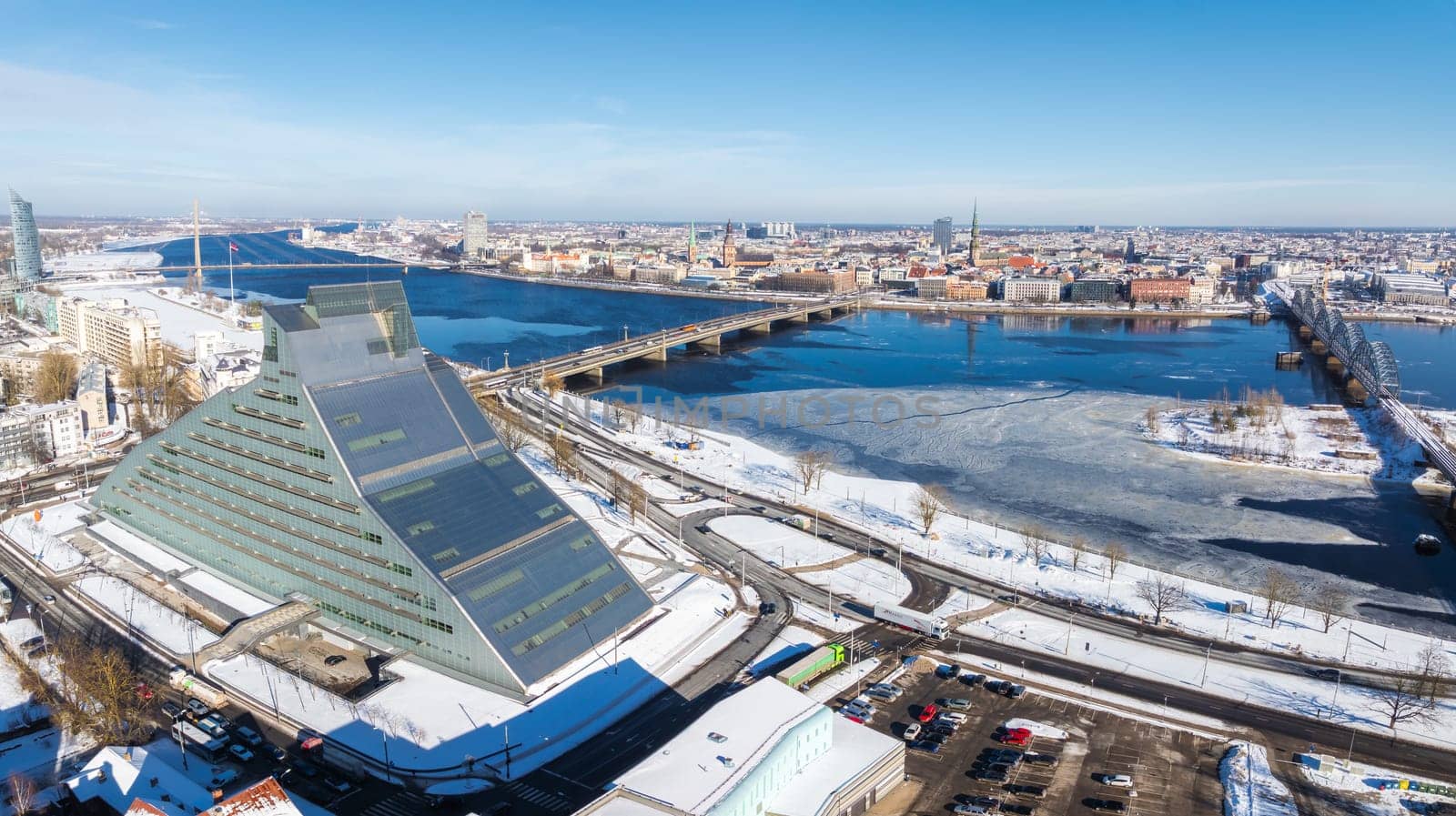 Aerial view of Riga, Latvia, featuring the National Library's modern architecture, the frozen Daugava River, and historic old town spires in winter.