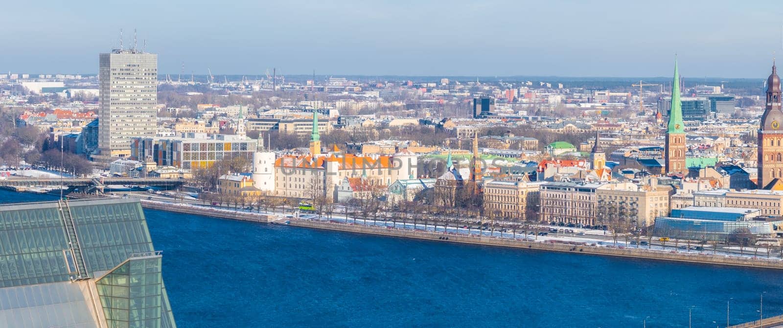 Aerial view of Riga, Latvia, in winter with the Daugava River, National Library, Riga Castle, Old Town spires, and snow dusted rooftops visible.