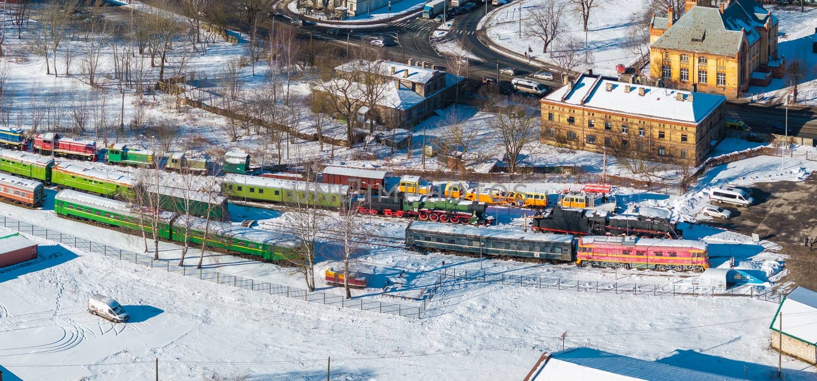 Aerial view of the Latvian Railway History Museum in Riga, featuring colorful vintage trains on snowy tracks, historic buildings, and bare winter trees.