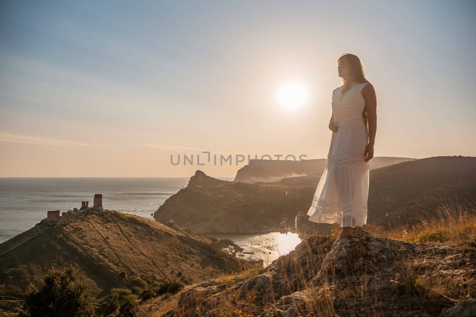 A woman stands on a rocky hill overlooking the ocean. She is wearing a white dress and she is enjoying the view. The scene is serene and peaceful, with the sun shining brightly in the background