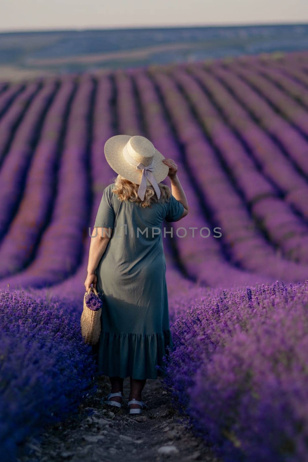 A woman wearing a straw hat stands in a field of purple flowers. She is holding a basket and she is enjoying the scenery