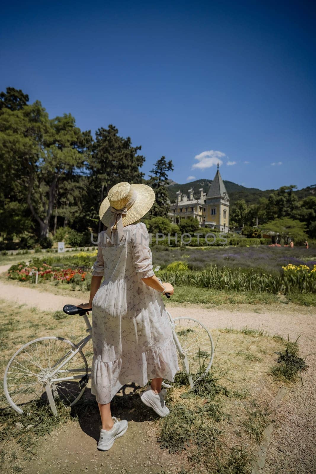 A woman in a white dress is standing next to a bicycle in a park. The scene is peaceful and serene, with a beautiful view of a house in the background. The woman is enjoying her time in the park