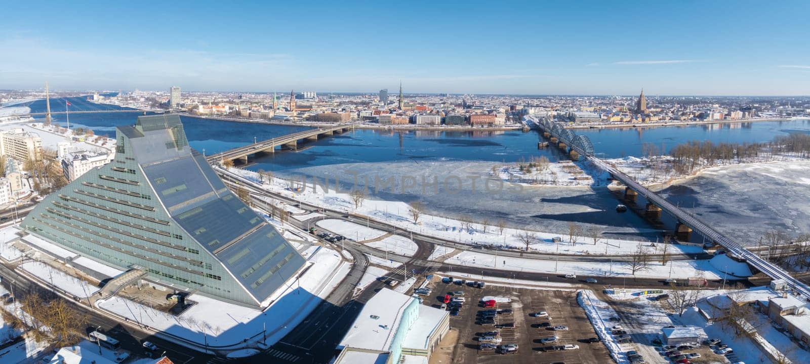 Aerial view of Riga, Latvia, featuring the National Library, Daugava River, Old Town spires, and Riga TV Tower under a blanket of snow.