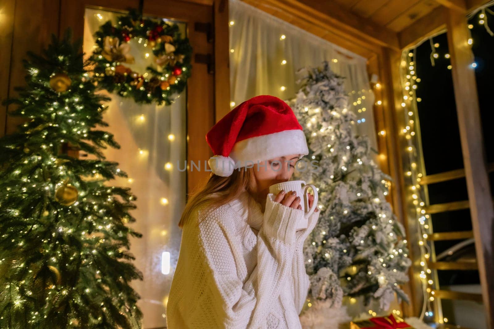 A woman wearing a Santa hat and white sweater holds a cup of coffee while standing in front of a decorated Christmas tree.