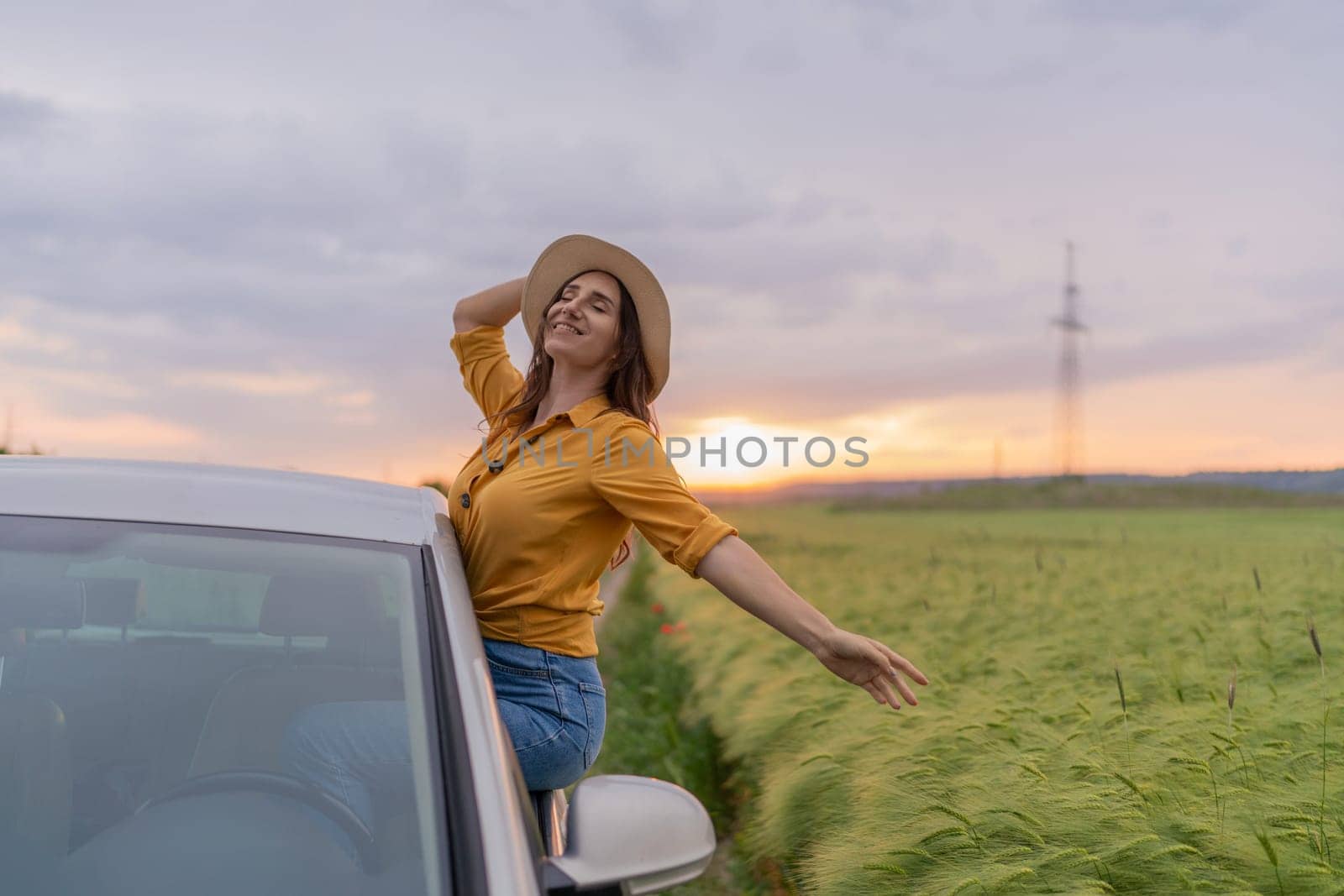 A woman is sitting in a car with her hat on and smiling. The sun is setting in the background