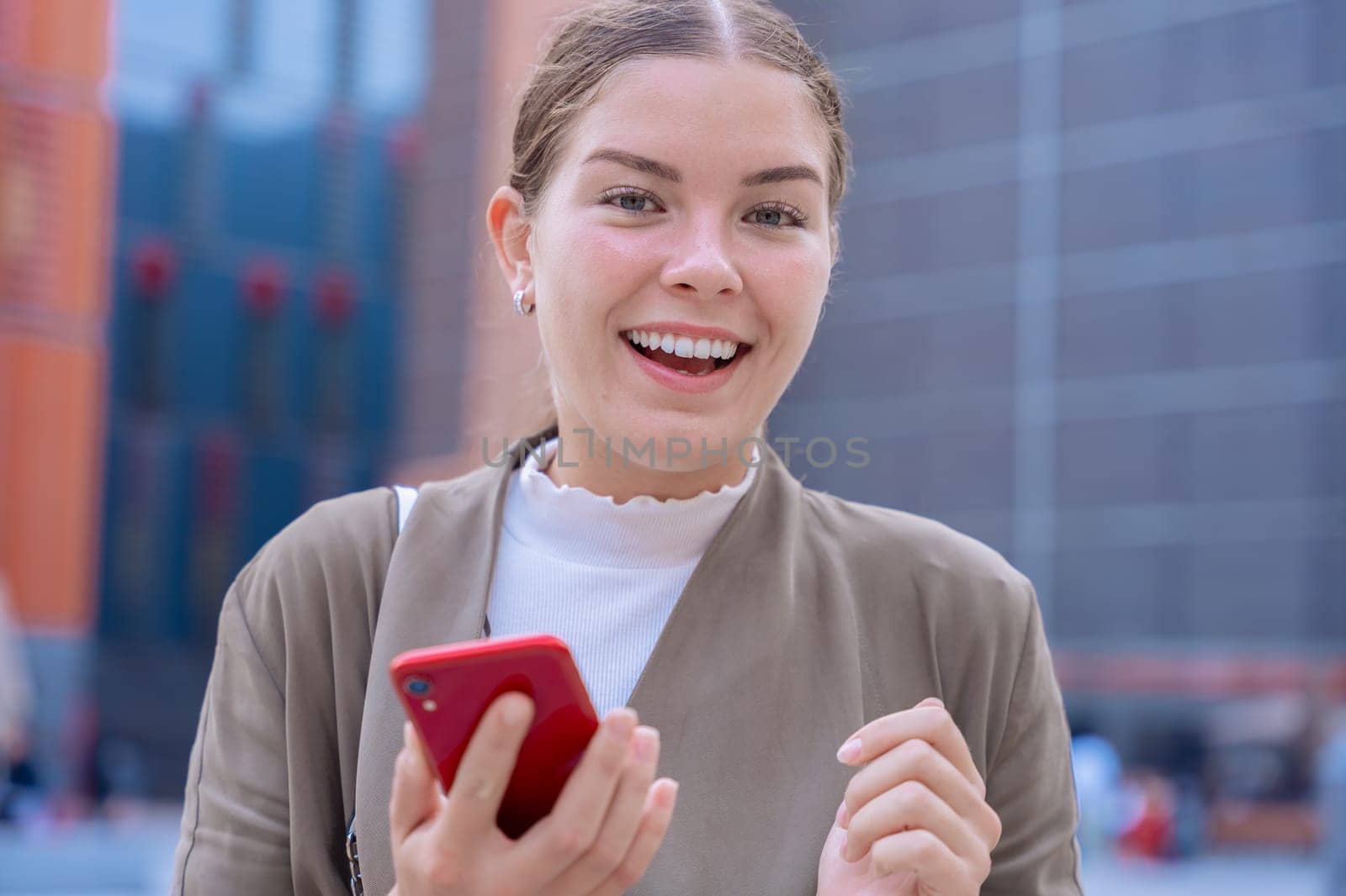 A Caucasian businesswoman wearing a blazer smiles as she checks her smartphone. Smiling, looking at the camera, standing, reacting to unexpected news or messages.