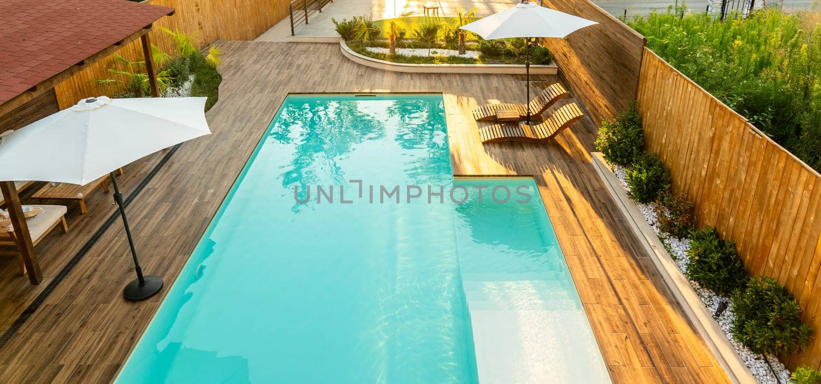Modern backyard with turquoise swimming pool, wooden deck, umbrellas, and lounge chairs in a serene setting