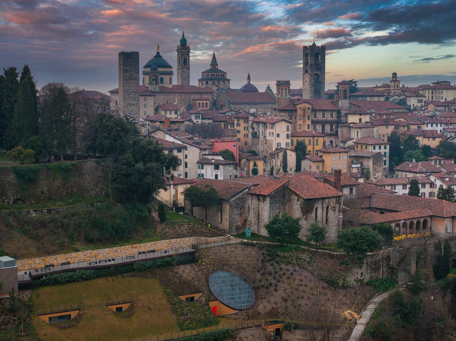 View of Bergamo's old town during sunset, featuring the Basilica of Santa Maria Maggiore, Cappella Colleoni, and Campanone, framed by gardens and greenery.