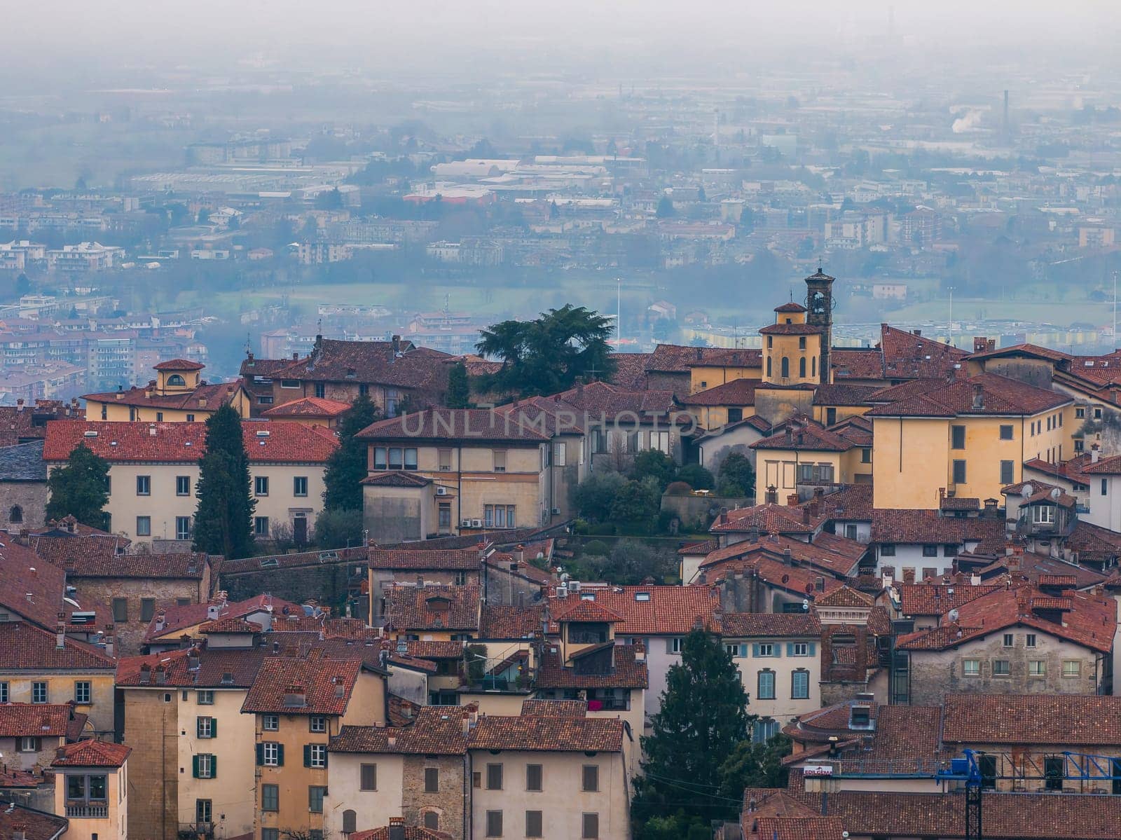 View of Bergamo's old town in Italy, featuring red tiled rooftops, a small bell tower, and a hazy backdrop of the modern city and countryside.