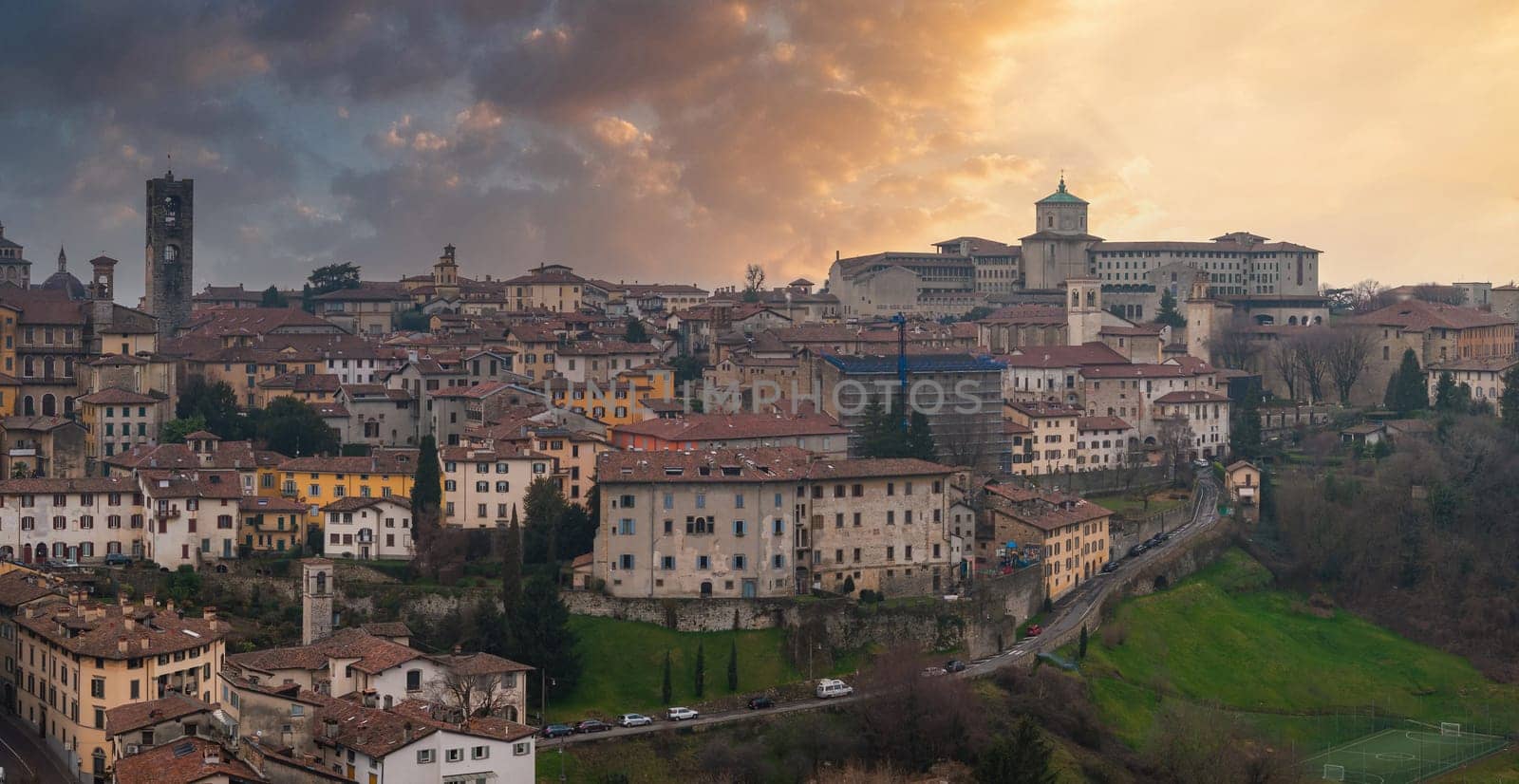 View of Bergamo's old town in Italy during sunset, featuring terracotta roofs, a bell tower, a green domed church, and a lush green hillside.