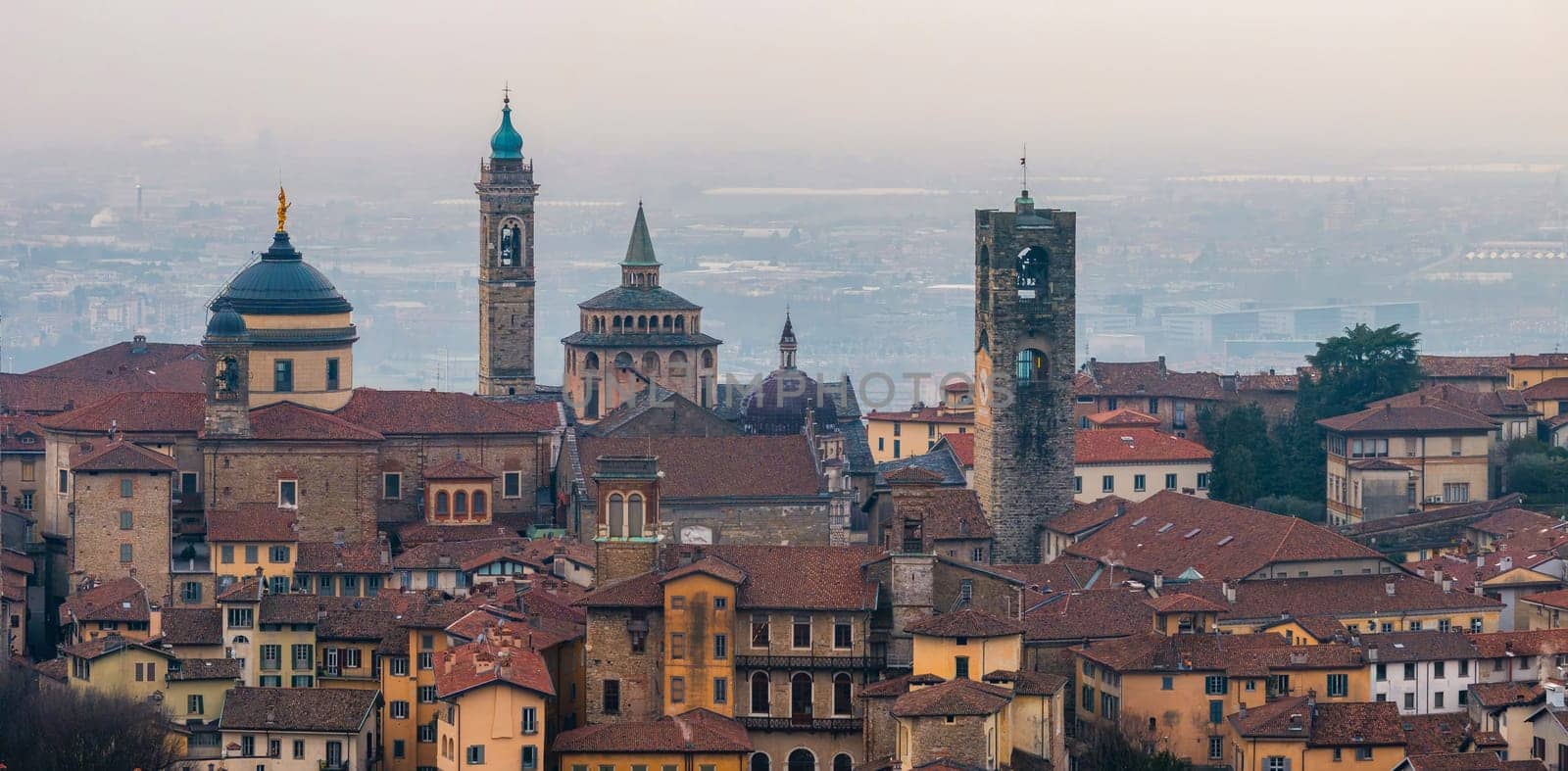 View of Bergamo's old town featuring the Basilica of Santa Maria Maggiore, Campanone, and Cappella Colleoni amid terracotta roofs and a hazy sunset.