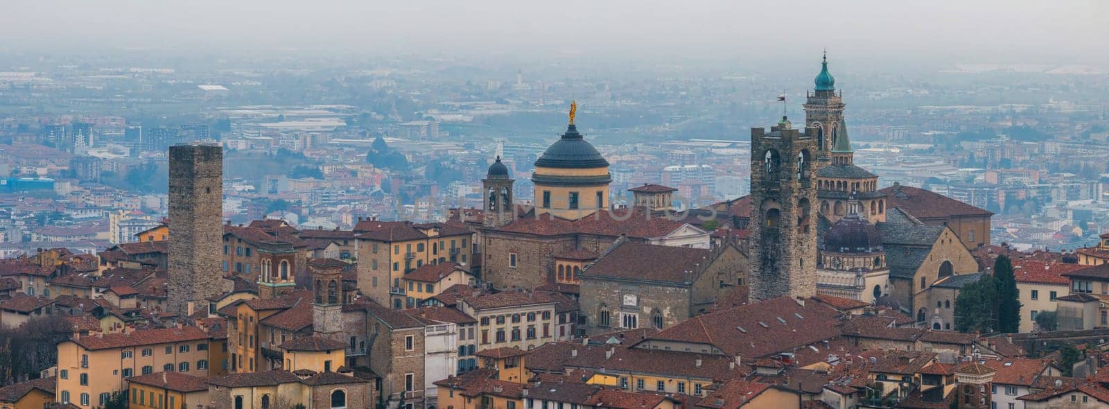 View of Bergamo's old town, featuring red tiled rooftops, the Basilica of Santa Maria Maggiore, Torre del Gombito, and a hazy modern city backdrop.