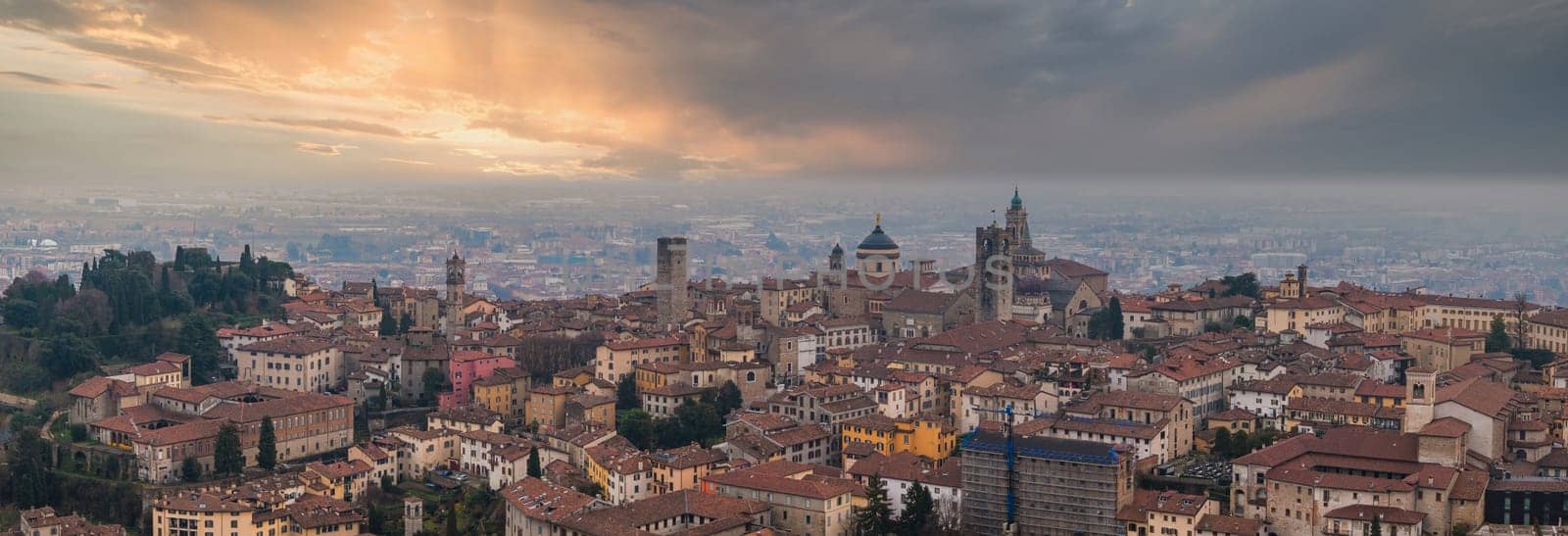 View of Bergamo's old town during sunset, featuring the Basilica of Santa Maria Maggiore, Torre del Campanone, terracotta rooftops, and dramatic clouds.