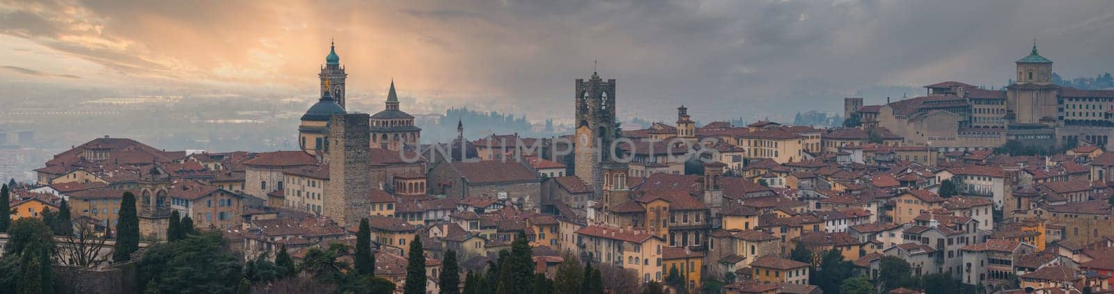 View of Bergamo's old town in Italy, featuring the Basilica of Santa Maria Maggiore, Torre del Gombito, red tiled roofs, and lush greenery at sunset.