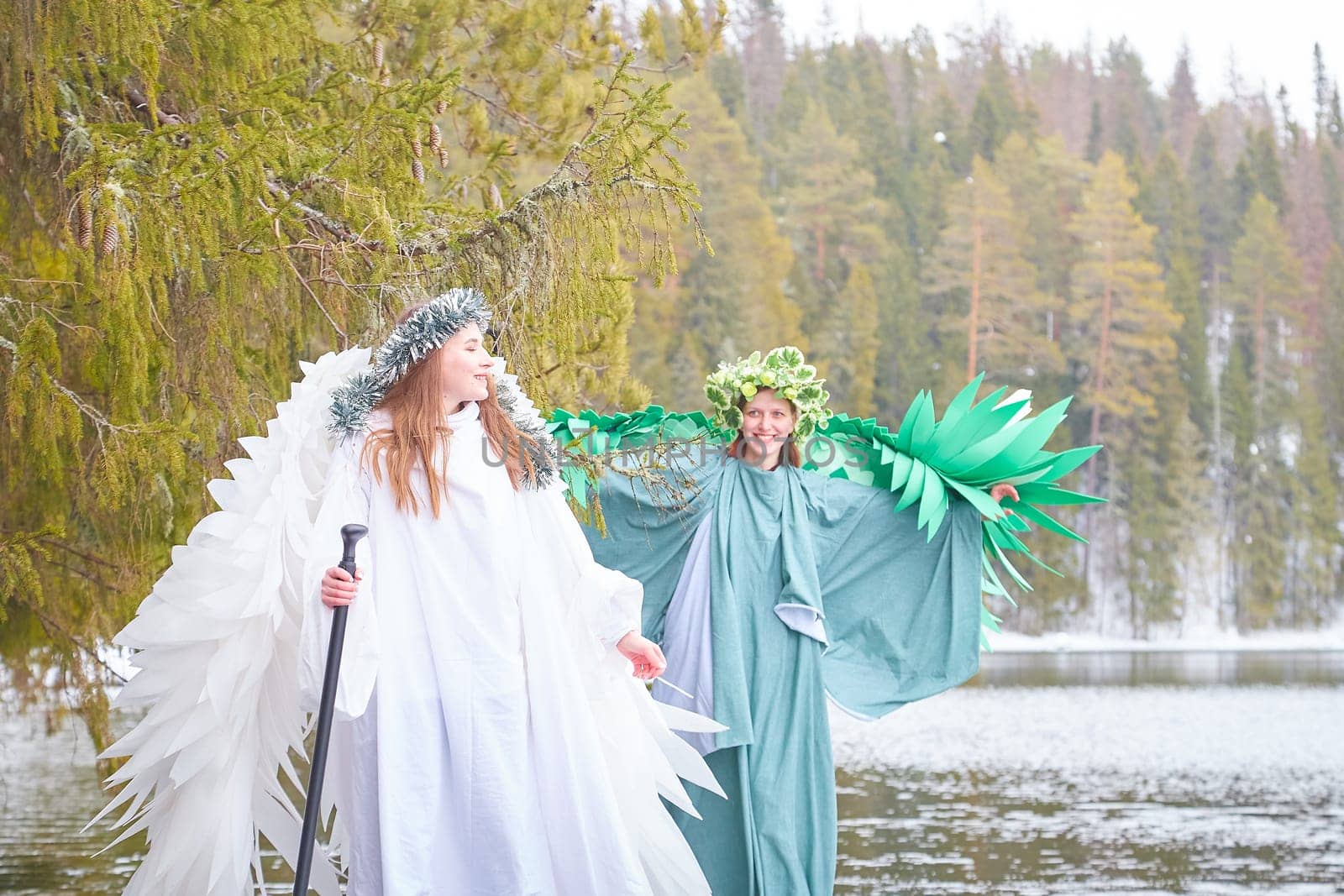 Two girls in flowing white and green dresses embody the transition between winter and spring near a serene lake, creating a fairy tale atmosphere.