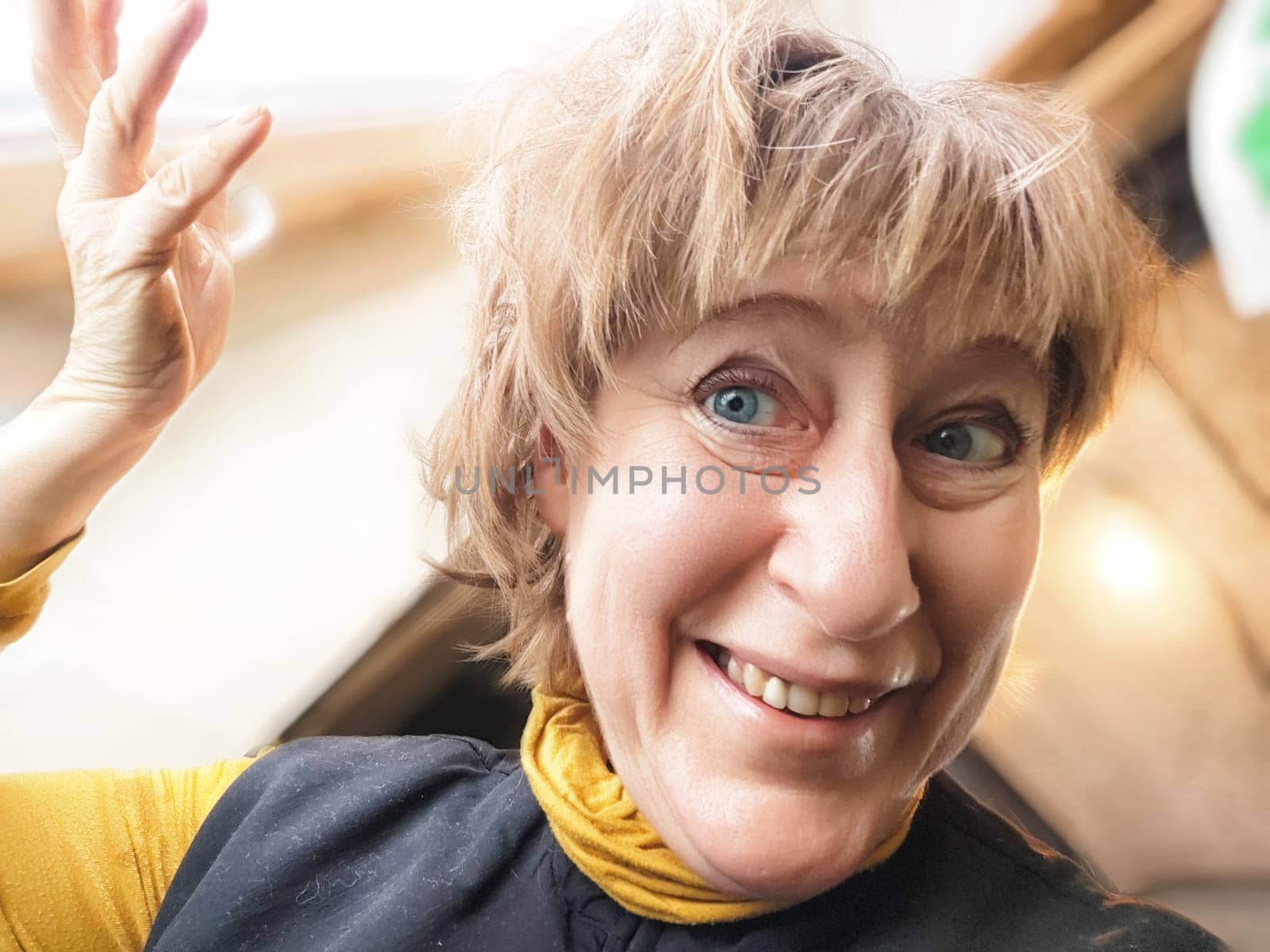 A middle-aged blonde woman smiles widely while taking a selfie indoors, sharing her vibrant travel experiences.