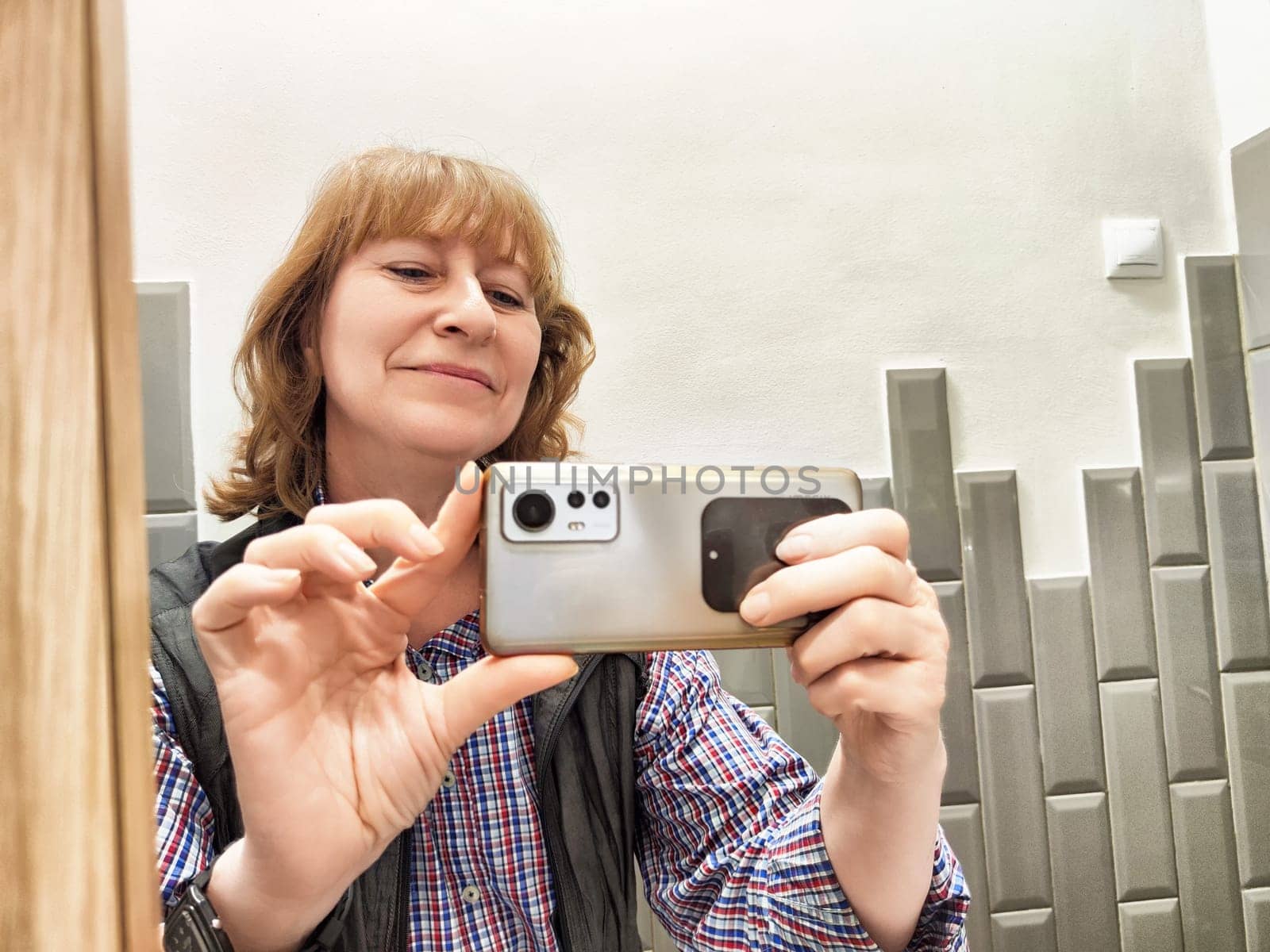 Middle-aged woman captures a cheerful mirror selfie while getting ready in a stylish bathroom during the morning.