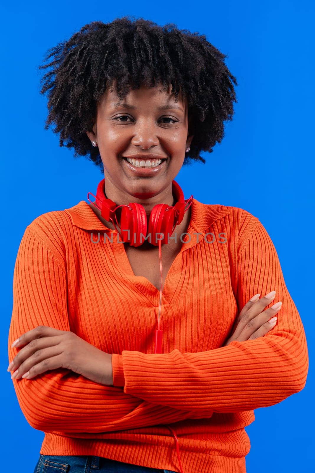 Smiling African American woman wearing red headphones,posing confidently with arms crossed against a blue background.Modern fashion, casual style, technology, self-assurance, and positivity.Copy space