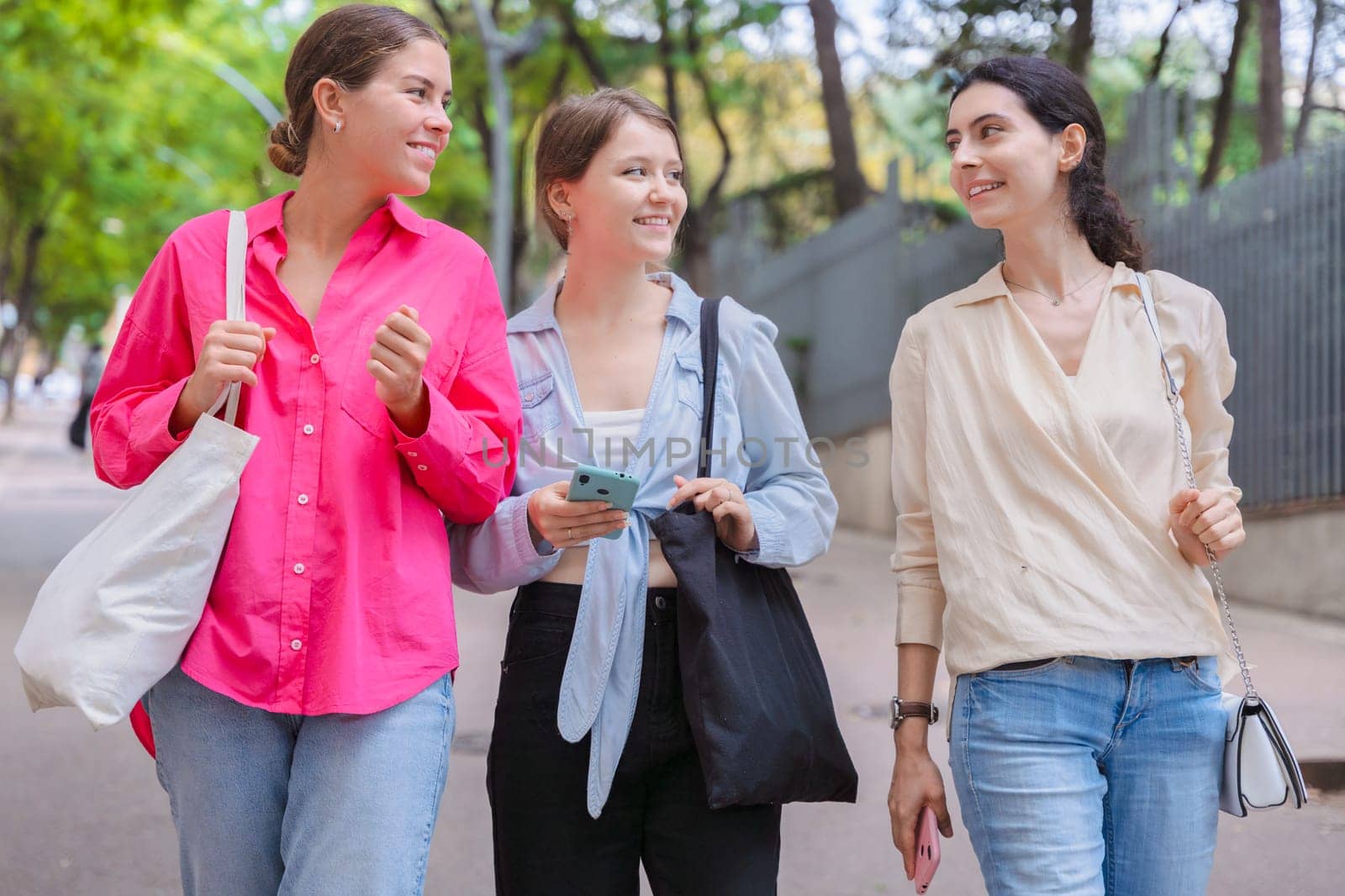 three gossipy teenagers chatting outdoors. A group of Caucasian friends chatting and communicating. Young adult students smiling and walking together at a social gathering.