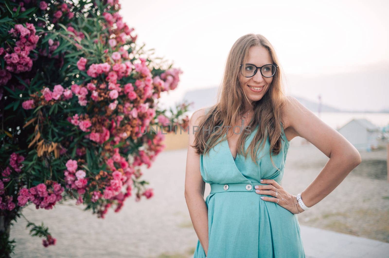 A graceful woman in a turquoise dress poses near blooming pink flowers, enjoying a peaceful moment in a natural outdoor setting.