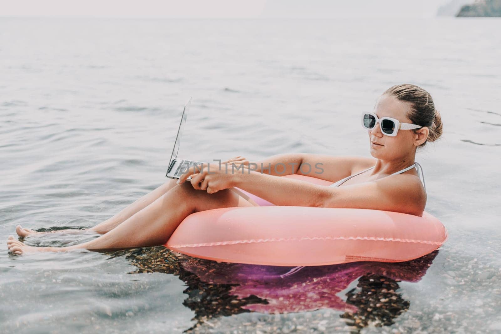 A woman is sitting in a pink inflatable raft on a lake, holding a laptop. Concept of relaxation and leisure, as the woman enjoys her time in the water while working on her laptop