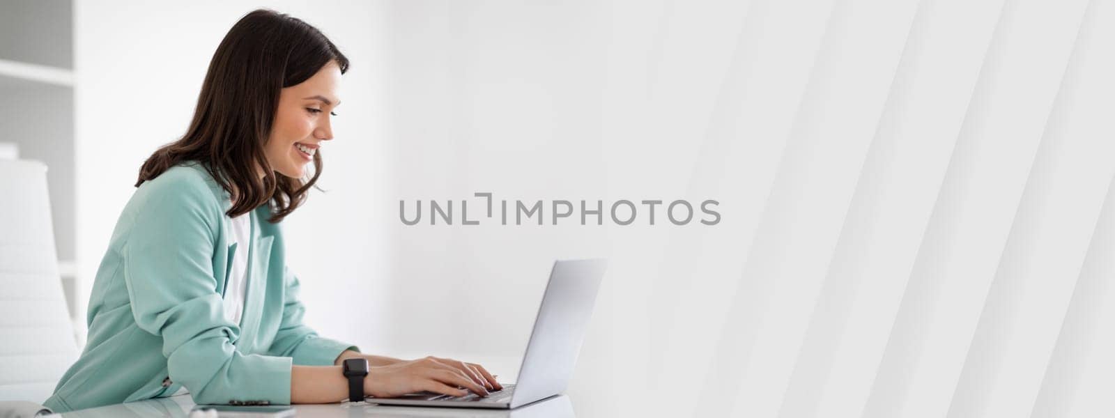 Smiling young european lady in suit at table typing on laptop, chatting with client in modern office interior, profile, ad, offer. Businesswoman and business, occupation, work, lifestyle with device