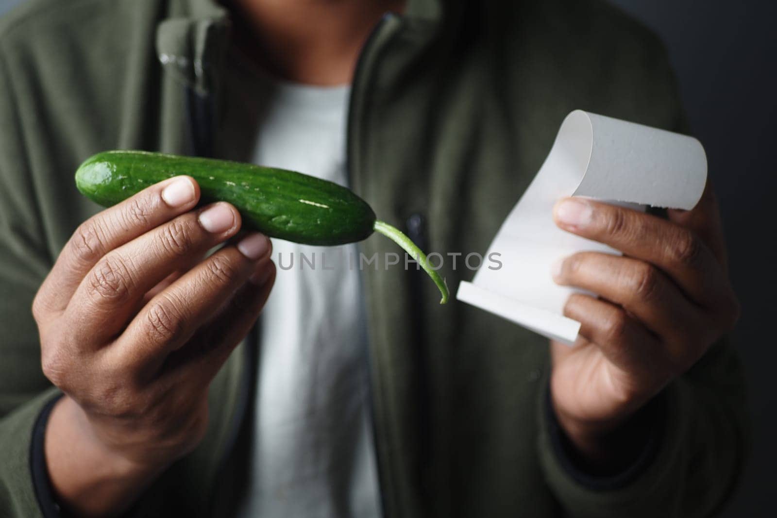 Man holds cucumber and receipt in indoor setting while examining them.