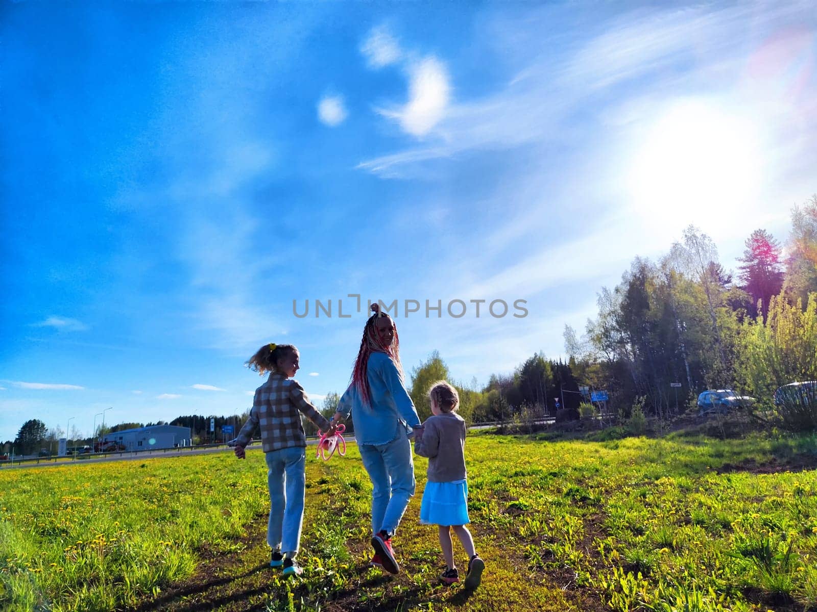 An adult and two children stroll together through a vibrant green field, enjoying a clear and sunny afternoon.