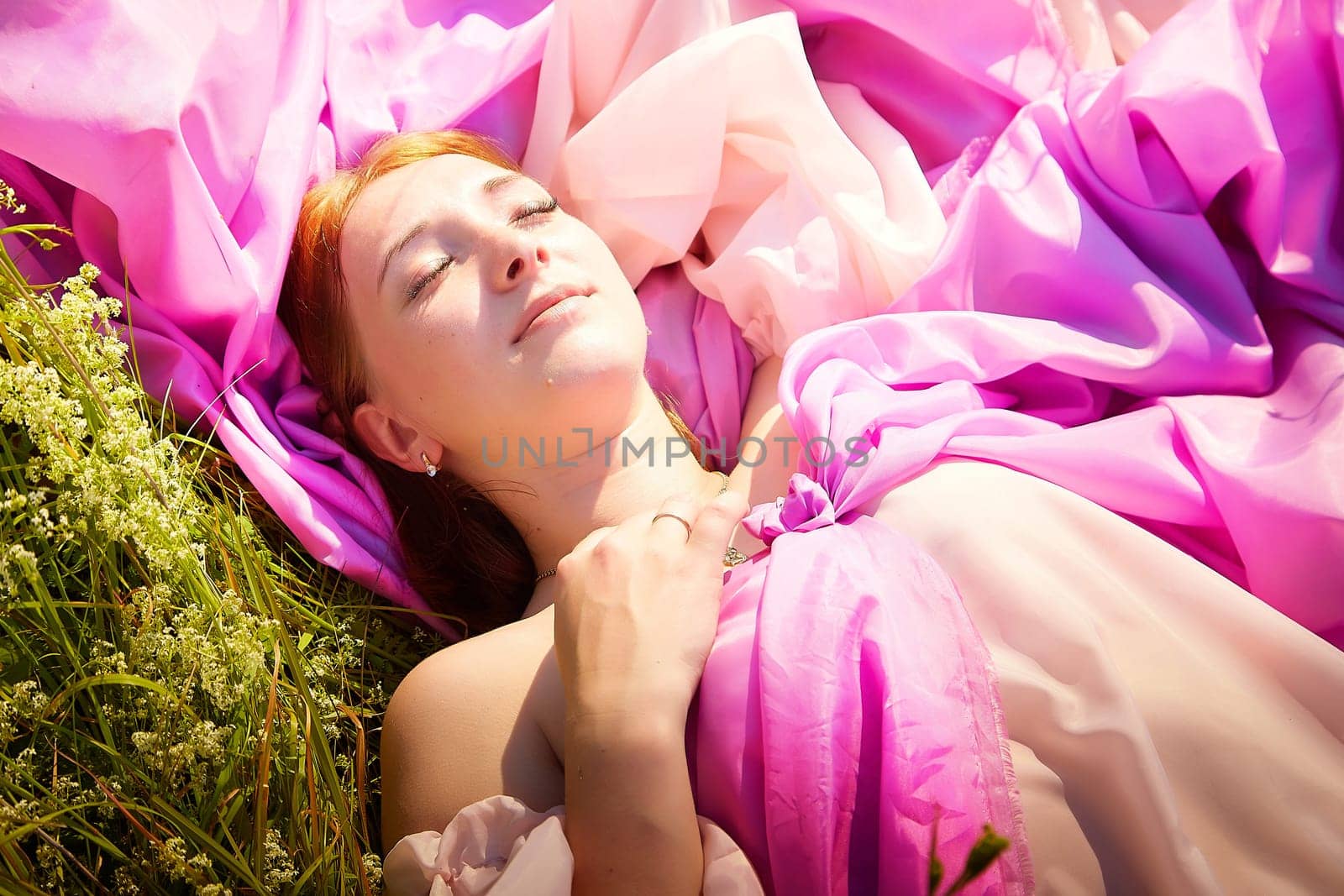 A woman enjoys a peaceful moment lying in a field, draped in bright pink fabric, absorbing the warm sunlight and nature around her.