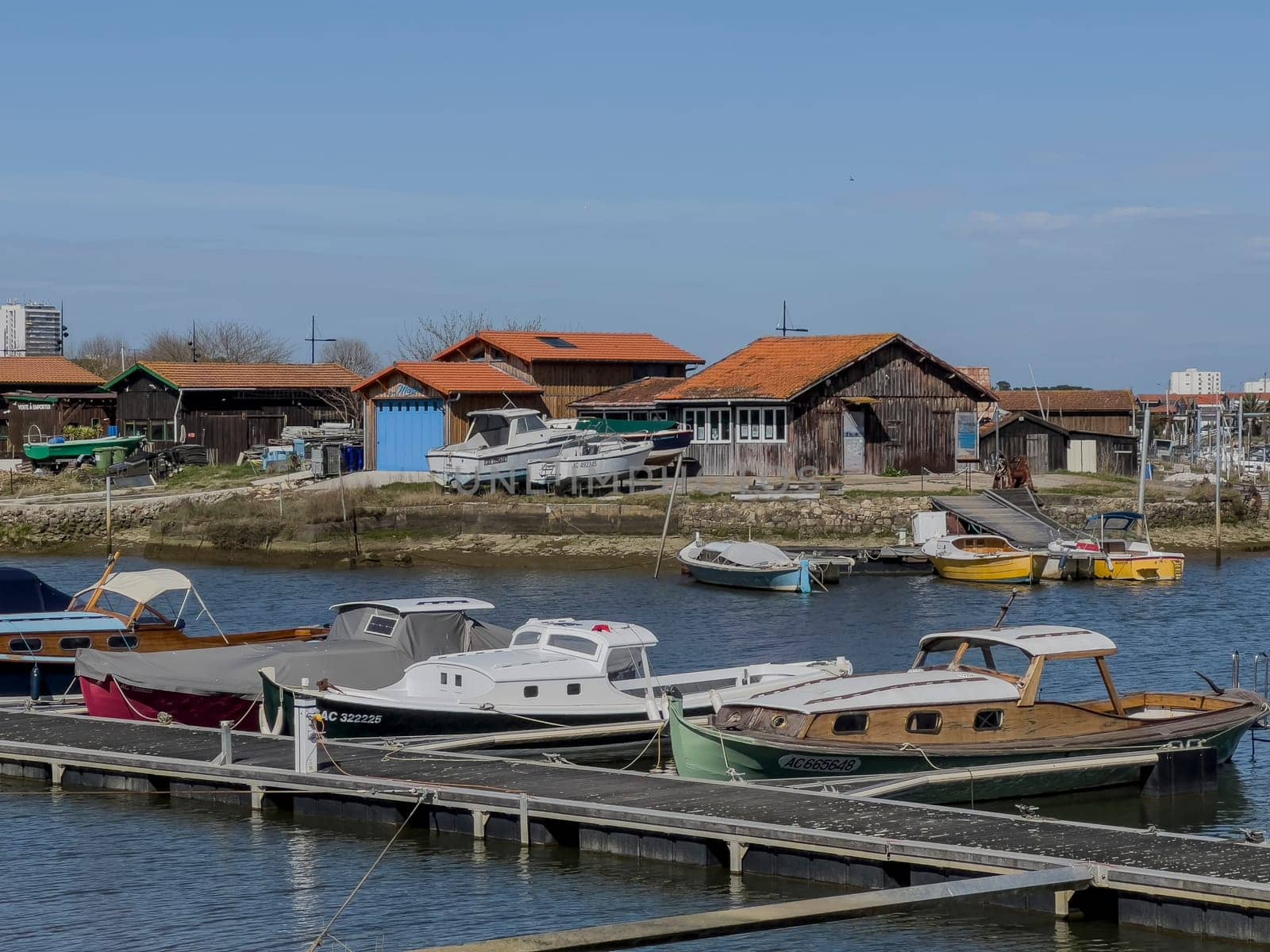 Boats line the dock in Port La Teste de Buch, showcasing various colors under a clear blue sky, while rustic buildings provide a charming backdrop to the tranquil waterfront setting.