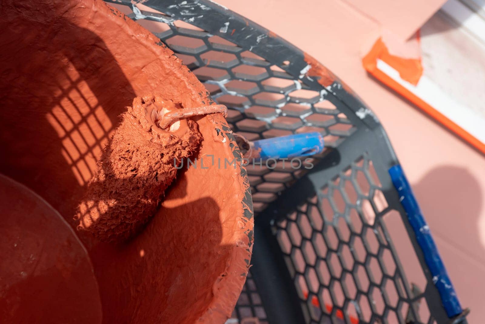 A paint roller rests in a bucket filled with reddish-brown paint. Surrounding tools and a work environment indicate ongoing home improvement activities in good weather.