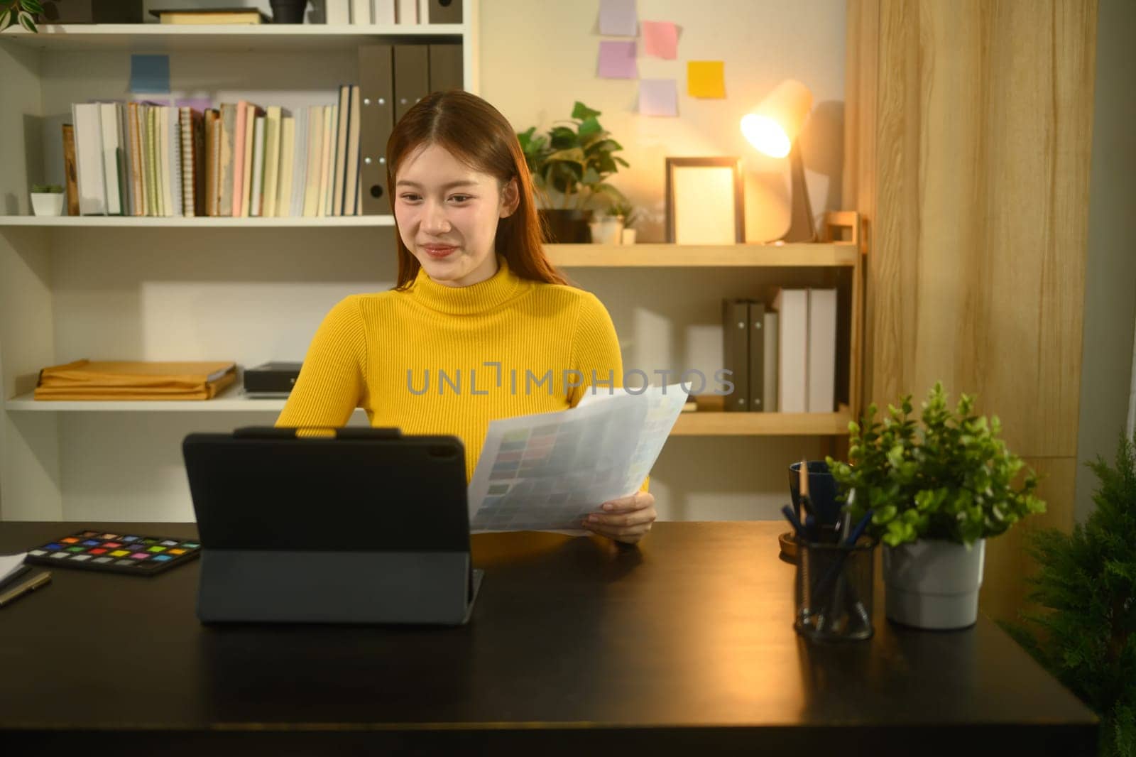 Young female graphic designer working on digital tablet and reviewing color samples at desk.