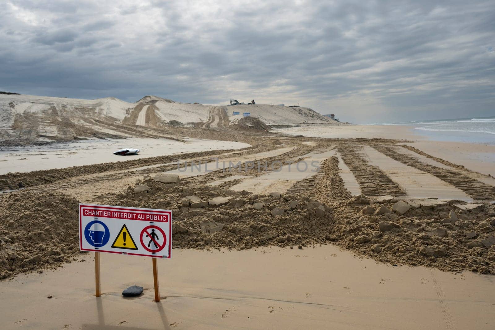 Signs indicate restricted access due to construction efforts aimed at managing coastal erosion while heavy machinery operates on the sand at Biscarosse beach under a cloudy sky.