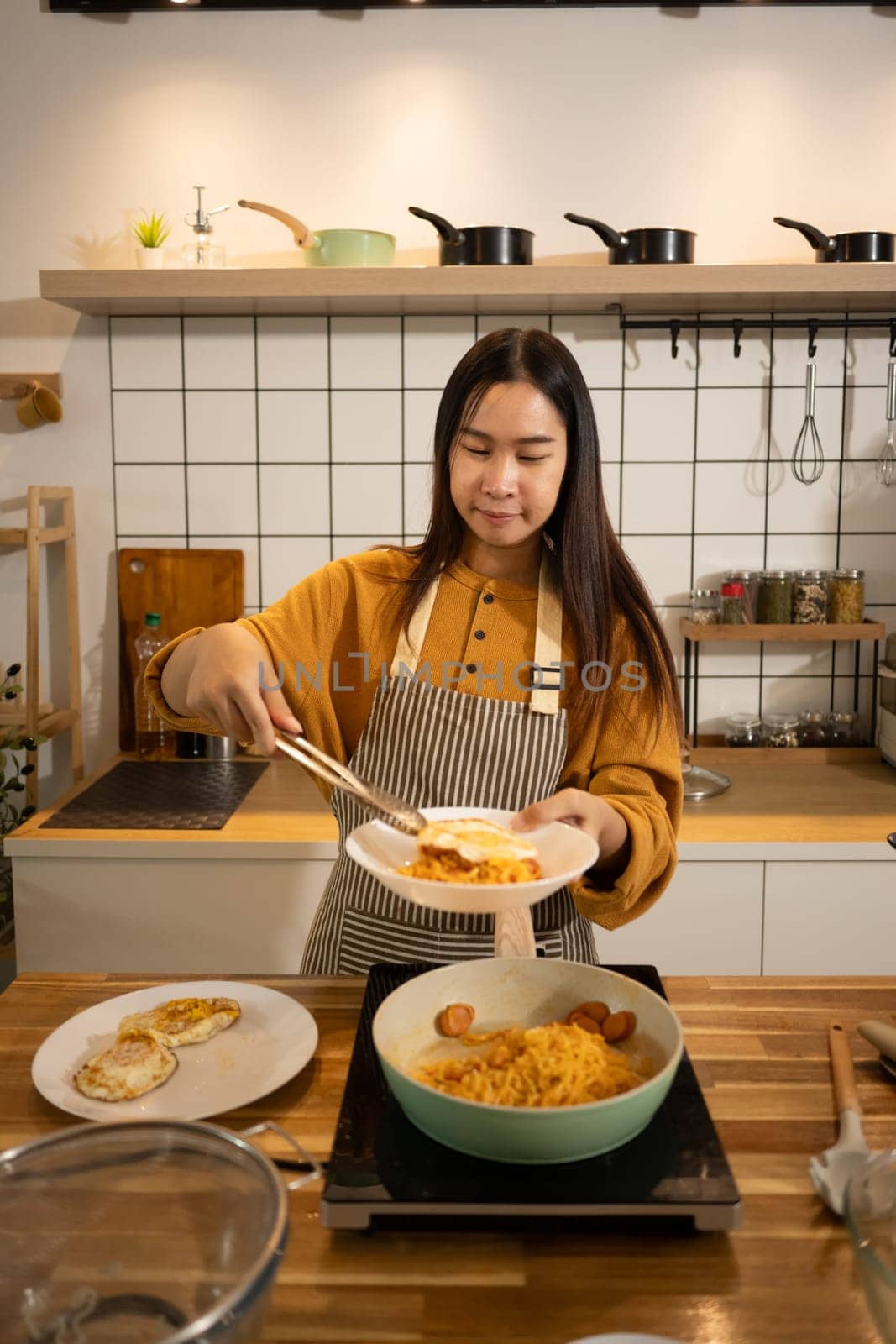 Young woman in an apron serving freshly cooked noodles onto a plate in a kitchen.