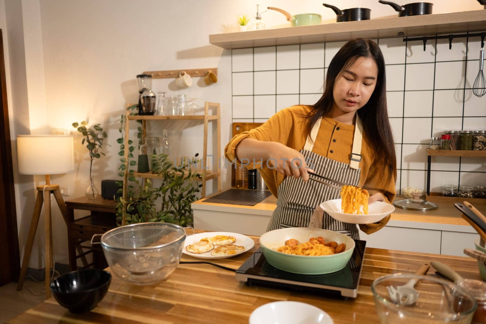 Young woman in an apron serving freshly cooked noodles onto a plate in a kitchen.