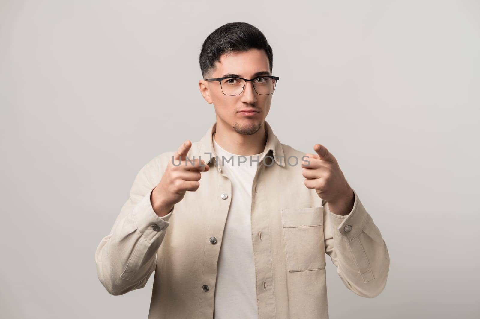 Confident man in beige clothes and eyeglasses points both fingers towards the camera with an encouraging smile, inspiring determination and self-belief in a supportive and motivating way.