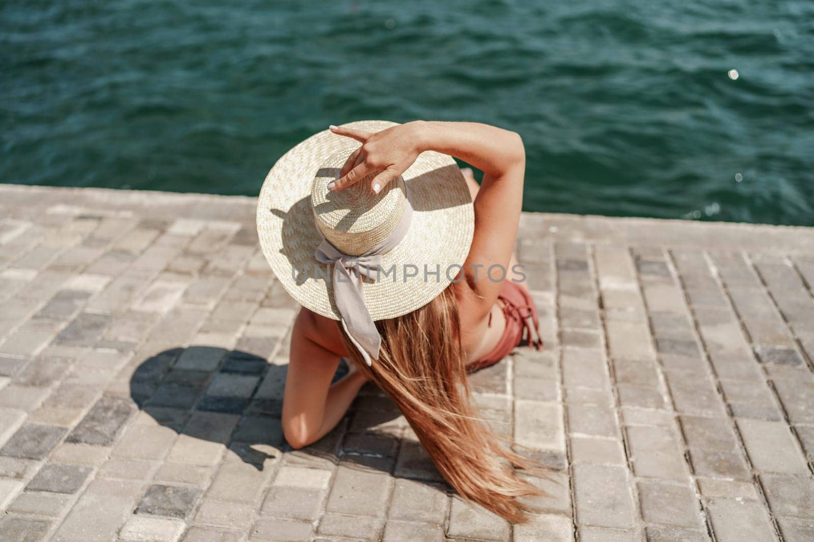 A woman in a swimsuit sits with her back holding a hat, looks at the ocean, sunny day, relaxes