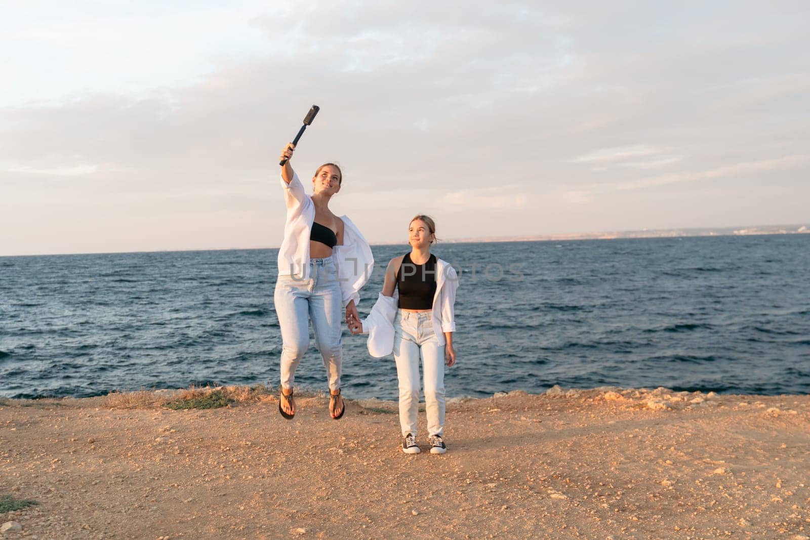 Two women in white shirts and jeans are jumping on a beach, one holding a wooden bat in the air. The ocean and a bright sky are in the background.