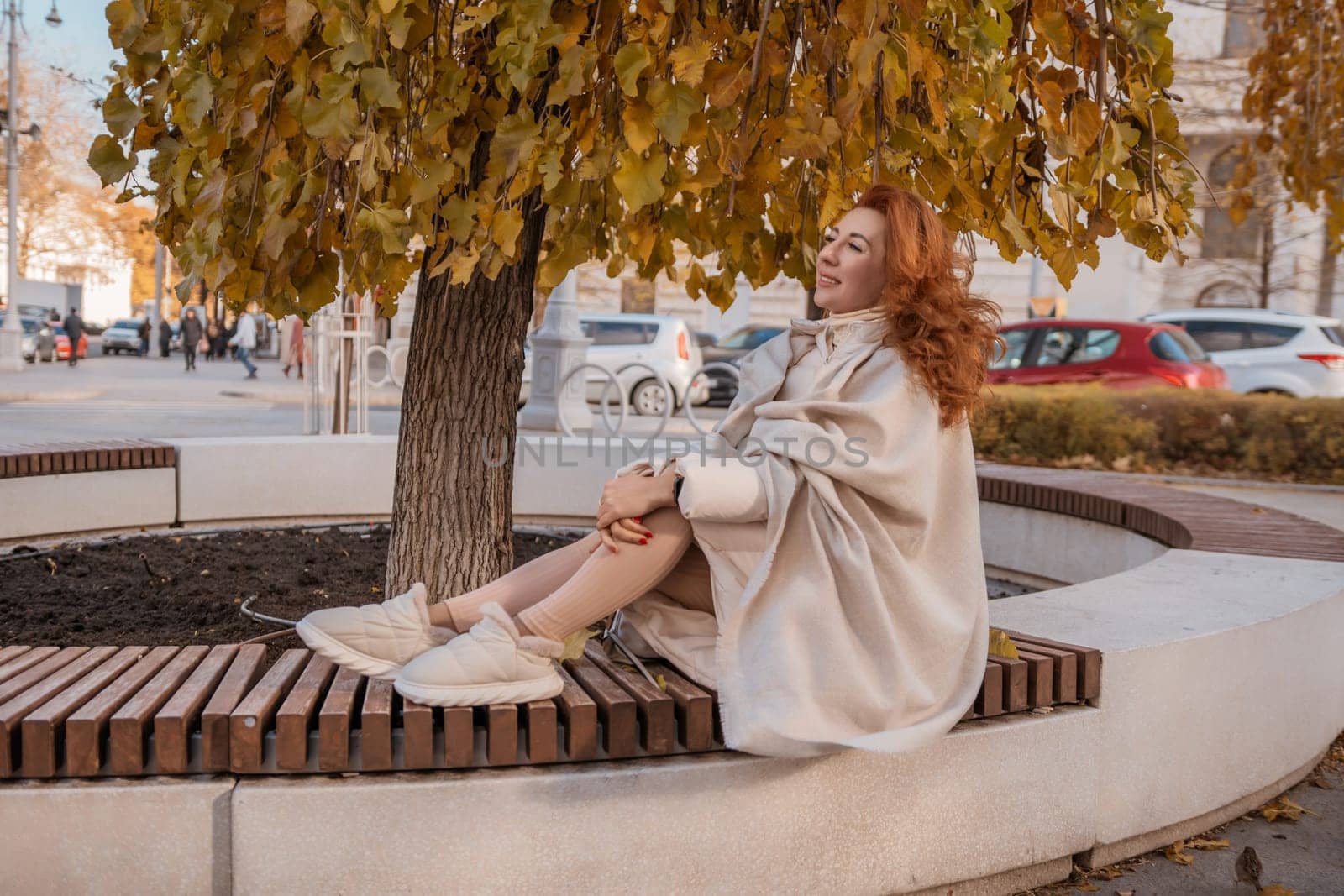 Redhead woman in beige coat sits on a park bench beneath an autumnal tree. She's wearing sneakers. City street background.