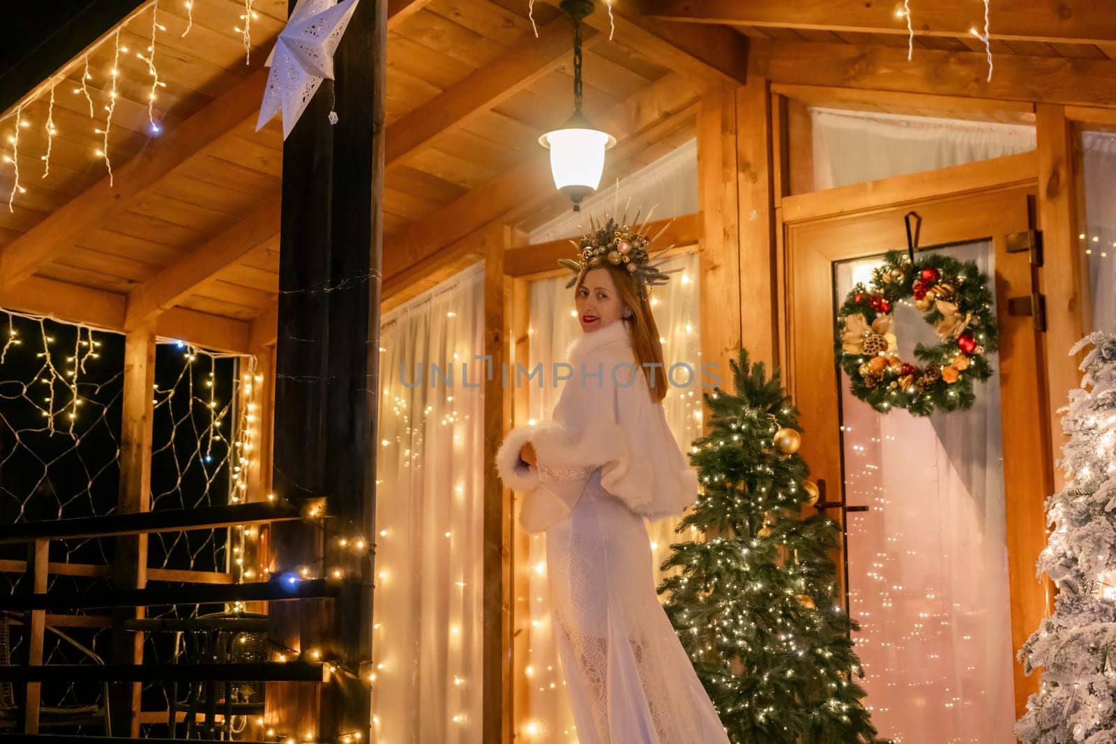 A woman in a white fur coat stands in front of a wooden cabin decorated with Christmas lights and a wreath.
