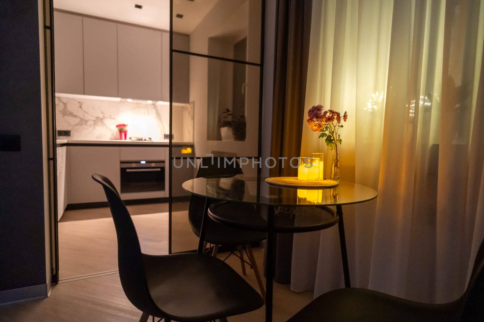 Modern apartment interior. Glass partition separates dining area with round table and chairs from a minimalist kitchen featuring marble countertops and built-in appliances. Warm evening lighting.