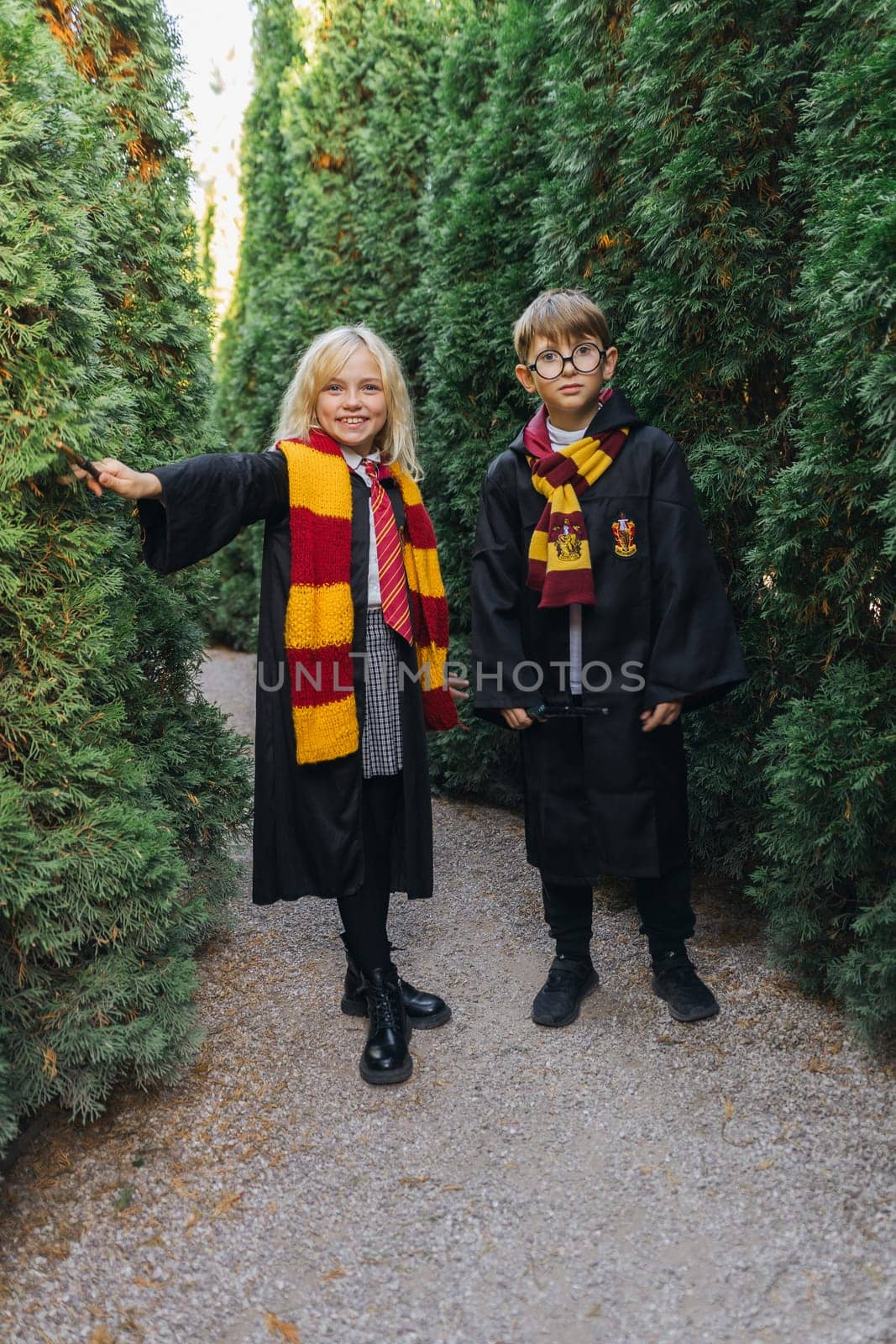 Two children dressed as Harry Potter characters stand in a hedge maze. The girl is on the left, her arm resting on a hedge. The boy is on the right, with his hands in his pockets.