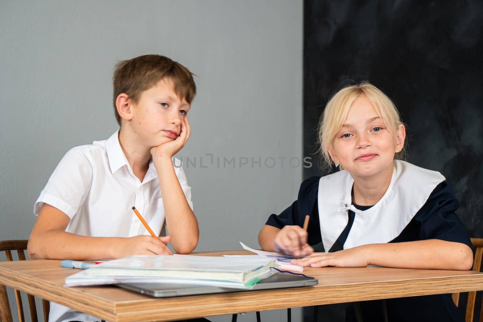 A boy and a girl in school uniforms sit at a desk writing in notebooks. The boy is looking away while the girl is smiling at the camera.