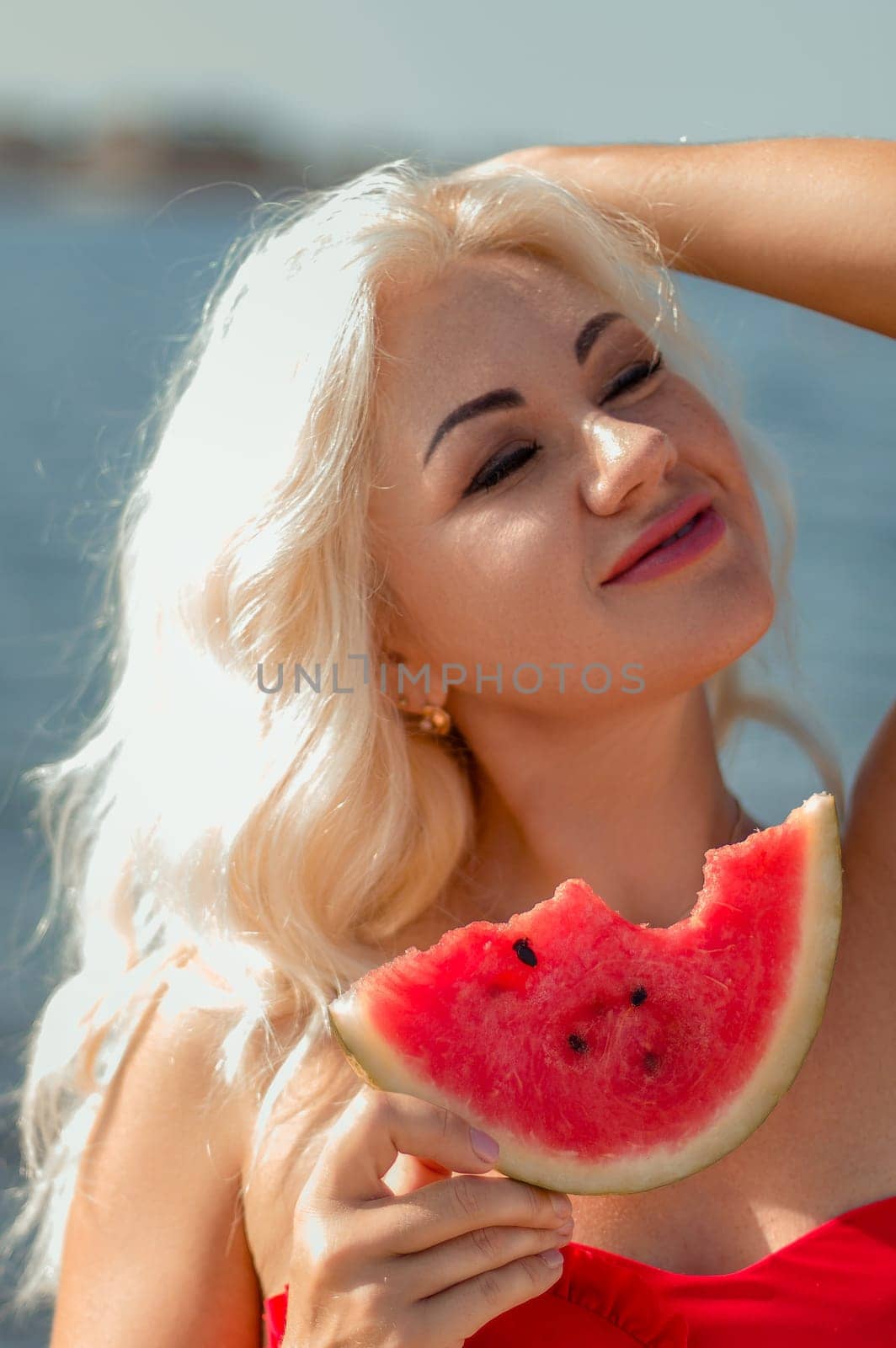 Blonde woman with long hair, wearing a red top, holds a slice of watermelon to her chest near the ocean. Sunlight illuminates her face.