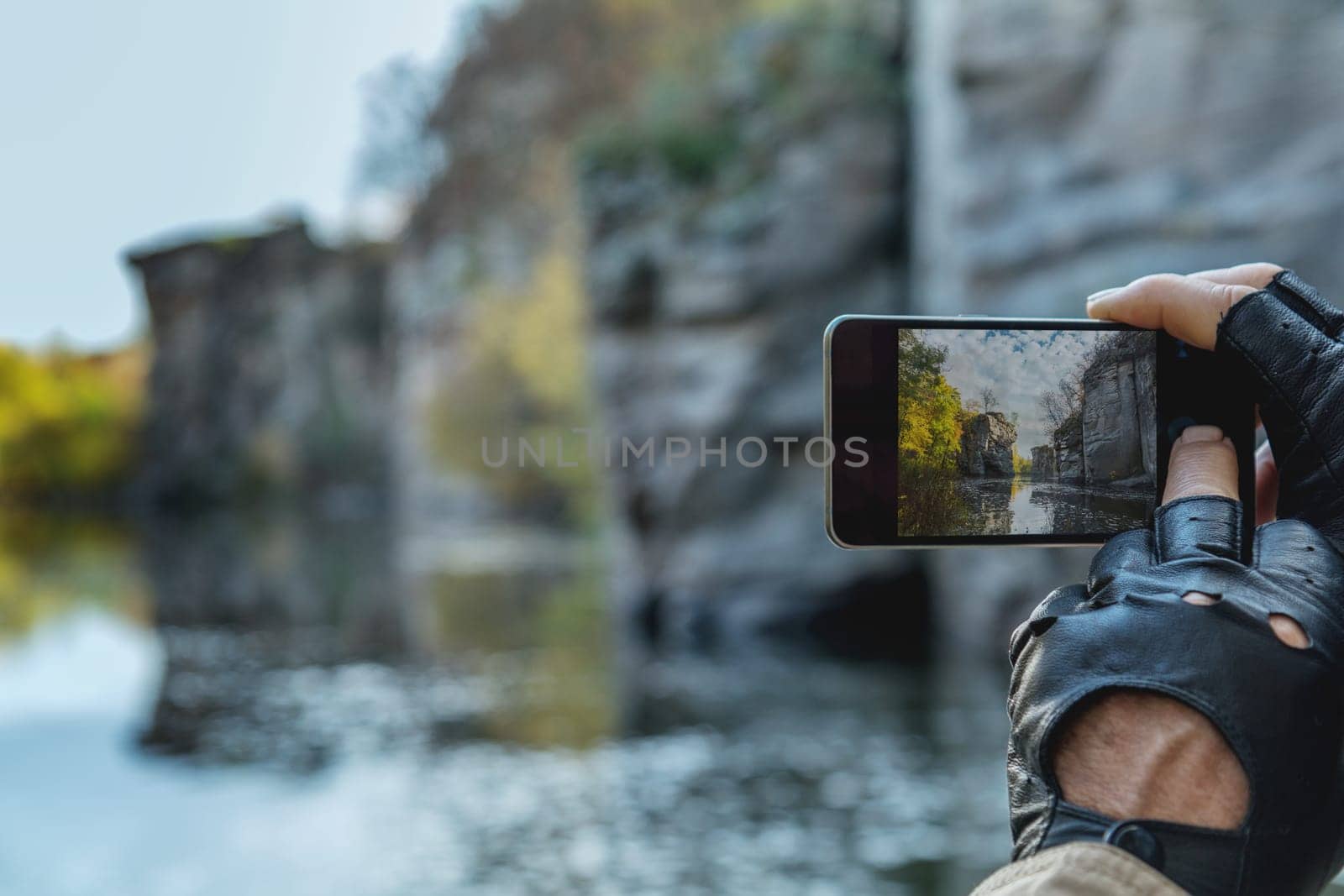 Photographer wearing black gloves taking pictures of a canyon landscape reflected in the water with a smartphone during autumn season