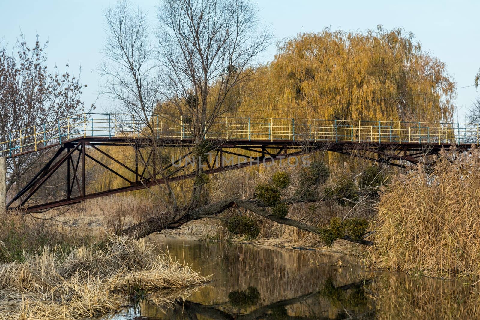Rusty metal bridge featuring yellow railing, reflecting in calm river water amidst a serene autumn landscape filled with colorful trees