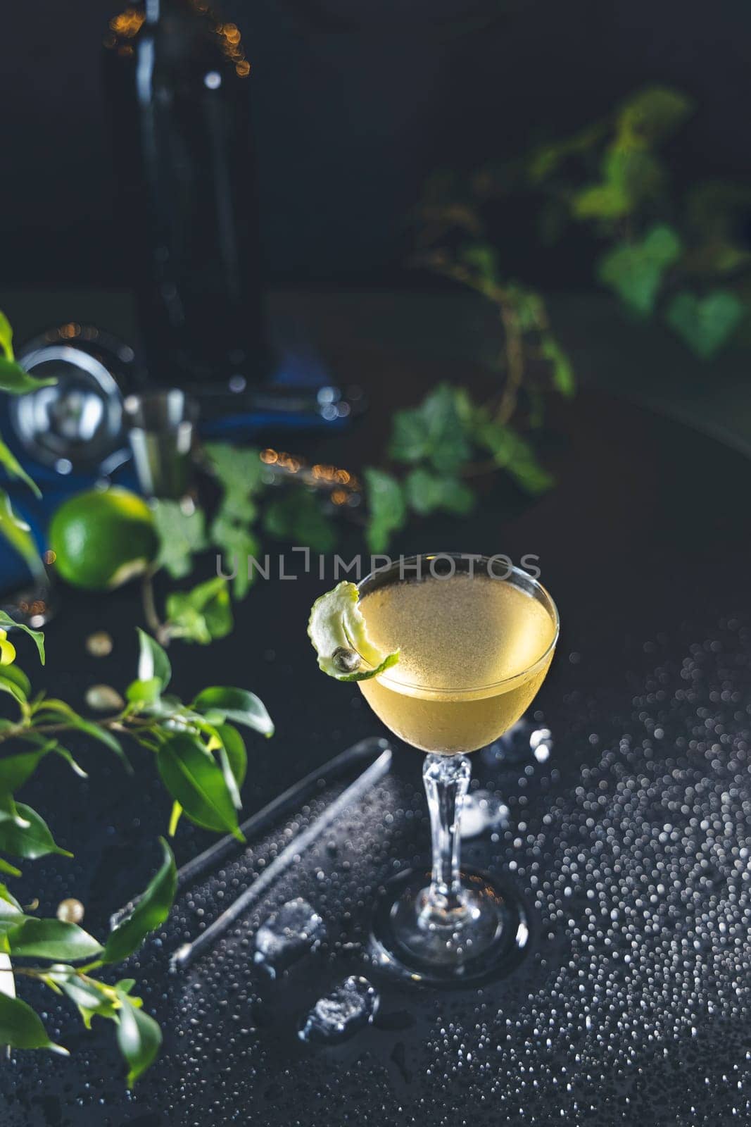 Refreshing cocktail served in a stemmed glass, garnished with a citrus twist, sits on a dark, wet bar top amidst ice and leafy greens