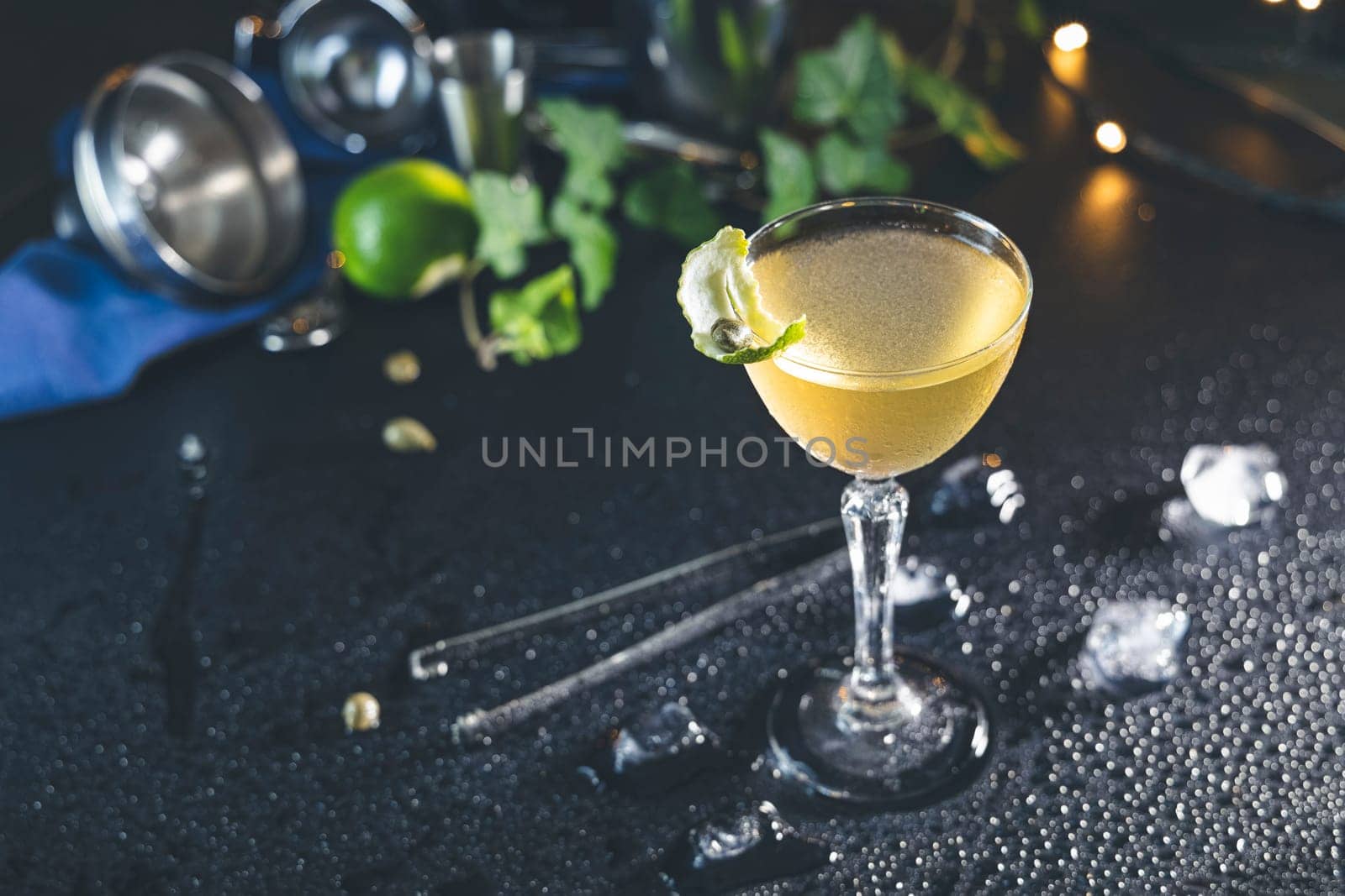 Refreshing daiquiri cocktail garnished with a lime peel, served in an elegant glass on a wet black table with bar equipment and ice cubes