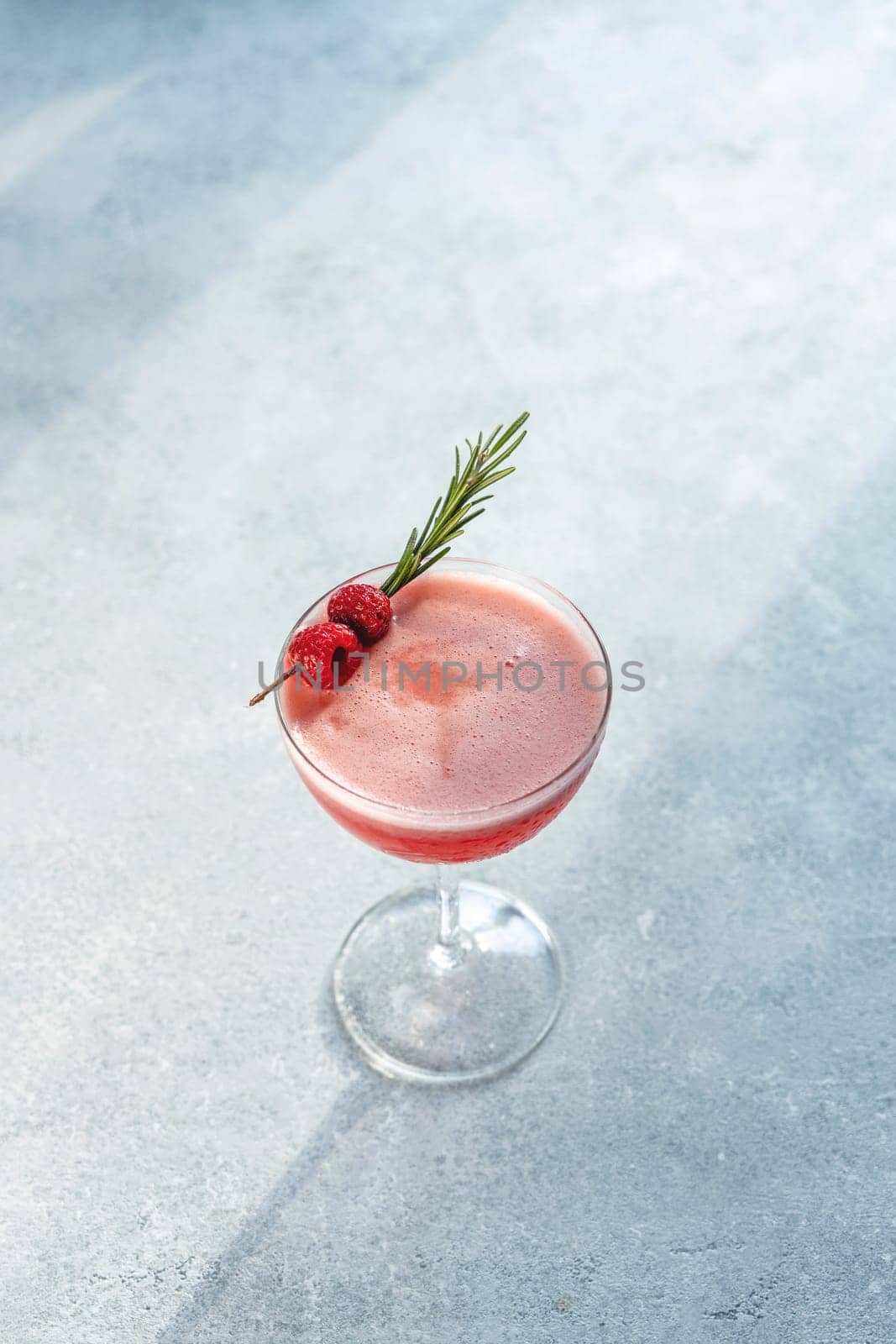 Coupe glass containing a vibrant pink cocktail garnished with fresh raspberries and a sprig of rosemary, creating an elegant and refreshing drink
